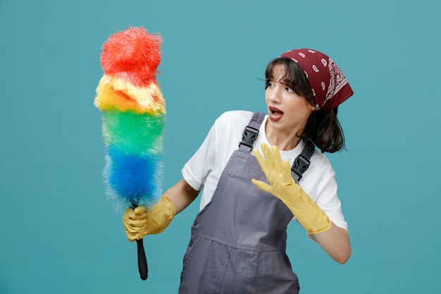 Scared young female cleaner wearing uniform bandana and rubber gloves holding and looking at feather duster showing empty hand isolated on blue background