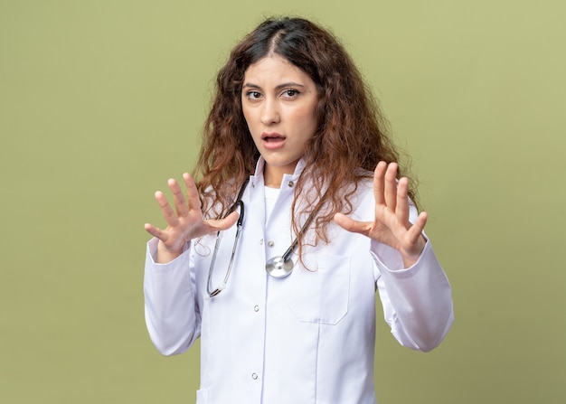 Scared young female doctor wearing medical robe and stethoscope doing refusal gesture isolated on olive green wall