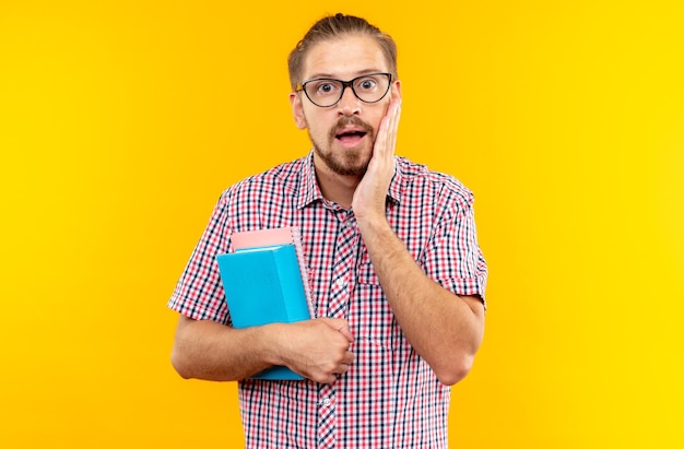 Scared young guy student wearing backpack with glasses holding book putting hand on cheek 