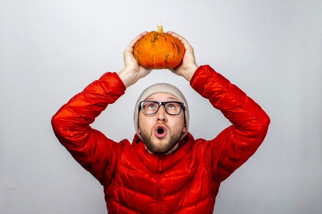 Photo scared young man in a red jacket and hat, holds a pumpkin over his head