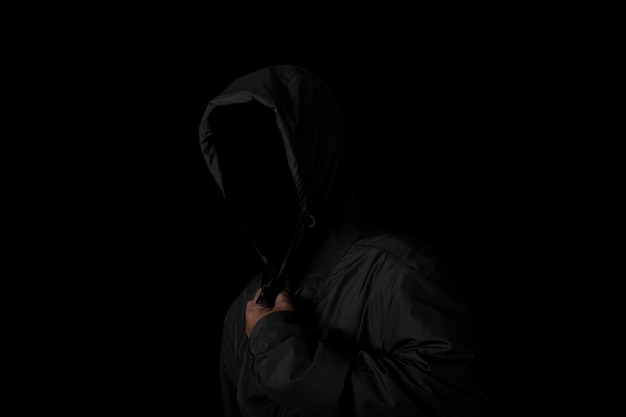 Страшный и жуткий человек, прячущийся в тени с лицом и личностью, скрытой под капюшоном Темный загадочный человек в толстовке с капюшоном на черном фоне