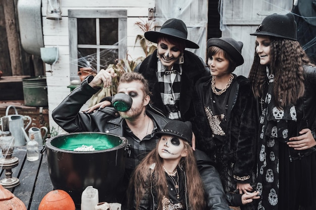 Foto spaventoso famiglia madre padre figlie che celebrano halloween calderone della pozioneteschio nero terrificante trucco a metà facciacostumi da stregaimmagini alla modaorroredivertimento alla festa per bambini nel fienile sulla strada