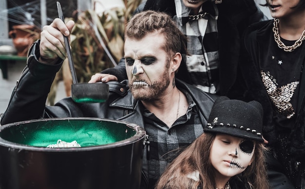 Scary family mother father daughters celebrating halloween Cauldron of potionTerrifying black skull halfface makeupwitch costumesstylish imagesHorrorfun at children's party in barn on street