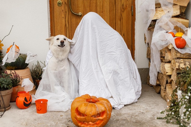 Scary ghost and cute dog with Jack o lantern at front of house with spooky halloween decorations on porch Trick or treat Person and puppy dressed as ghost trick or treating Happy Halloween