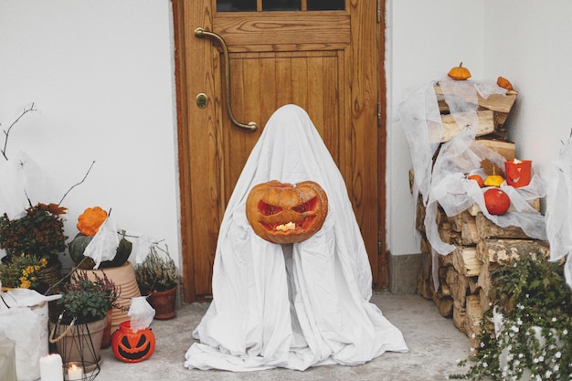 Scary ghost holding Jack o lantern at front of house with spiders web pumpkin and candle Trick or treat Spooky halloween decorations and kid dressed as ghost on porch Happy Halloween