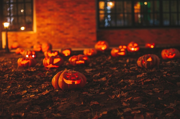 Scary jack lantern halloween pumpkins in darkness on ground among dry leaves at street Hallows eve decoration funny glow pumpkin with candles on dark background in open air near house