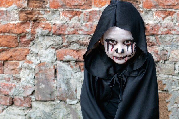 Scary little girl in a little zombie costume looks frighteningly at the camera against a brick wall.