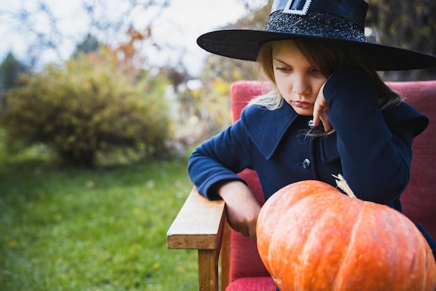 Scary little girl in witch costume hat with big pumpkin celebrating halloween holiday Sitting on armchair in coat with pumpkin Stylish image Horror fun at children's party in barn on street