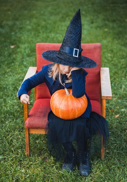 Scary little girl in witch costume hat with big pumpkin celebrating halloween holiday Sitting on armchair in coat with pumpkin Stylish image Horror fun at children's party in barn on street