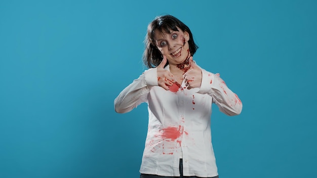 Scary looking monster showing thumbs up hand gesture on blue background. Approving spooky brain-eating walking dead corpse with deep and bloody wounds showing approval hand sign. Studio shot