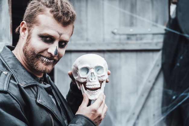 Scary man with dreadlocks looking at skull Celebrating halloween Terrifying black white face monster makeup and stylish costume imageHorrorfun at children's party in barn on streetShirtjackets