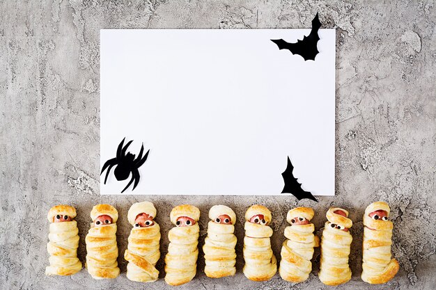 Photo scary sausage mummies in dough with funny eyes on table. halloween decoration and white blank paper note or greeting card