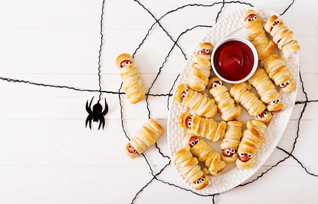 Scary sausage mummies in dough with funny eyes on table. Halloween food. 