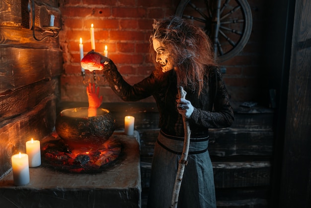 Scary witch reads spell over the pot with human body parts, dark powers of witchcraft, spiritual seance with candles.