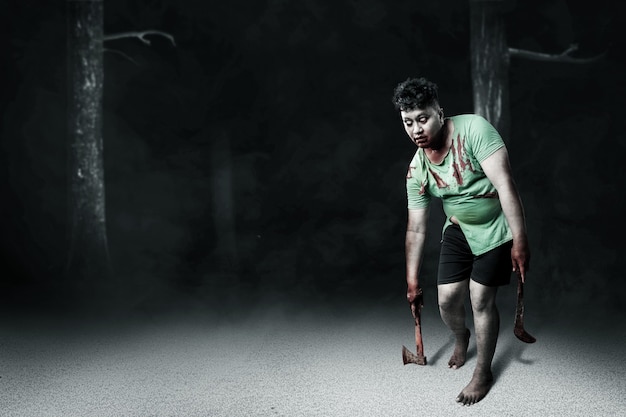 Photo scary zombie with blood and wound on his body holding ax and sickle walking with dark background