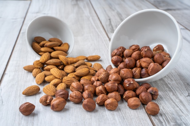 Scattered hazlenuts and almond seeds with white bowls on lignt wood table.