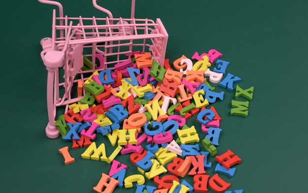 Photo scattered multicolored letters of the english alphabet from a miniature shopping cart on a green background, close up