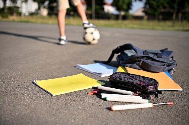 写真 開いたバックパックから落ちてくる散らばった学用品やワークブックは、サッカーボールの少年の足を背景に校庭のアスファルトの上に横たわっています。