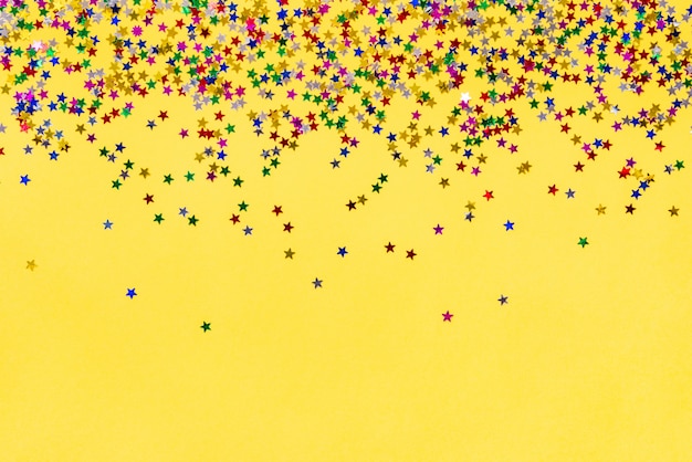 Scattered star shaped colorful glittering confetti on yellow background. Copy space.