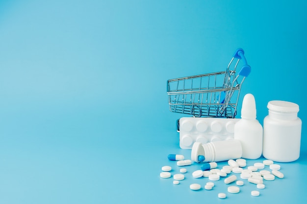 Scattered variety pills, drugs, spay, bottles, thermometer, syringe and empty shopping trolley cart on blue background