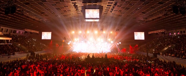 Scene illuminated by beautiful rays of lighting equipment. the\
concert crowd having fun at the center in the big hall. television\
is broadcast live. many people look towards the stage.