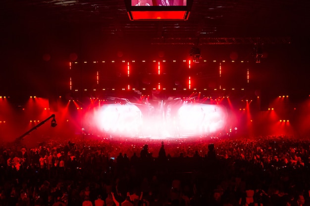 Scene illuminated by beautiful rays of lighting equipment. The concert crowd having fun at the center in the big hall. Television is broadcast live. Many people look towards the stage.