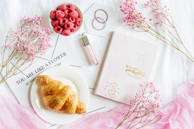 Scene with croissant pastry, fresh raspberries, notebook, lipgloss and pink flowers. 