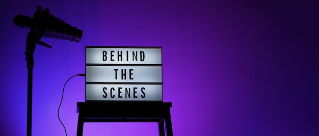 Behind the scenes text on Lightbox or Cinema Light box. Movie board light box on studio stair and light Snoot tripod on colors gradiant Background. Represent behind the scenes team is working on.