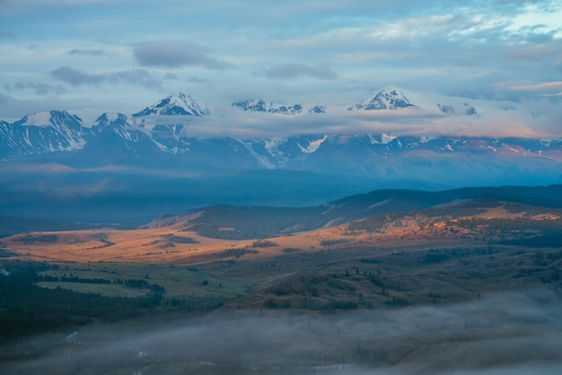 Живописный альпийский пейзаж с низкими облаками на обширном плато с горной рекой и лесом в солнечном свете на фоне великих заснеженных гор. Красочная горная долина и заснеженные горы с низкими облаками.