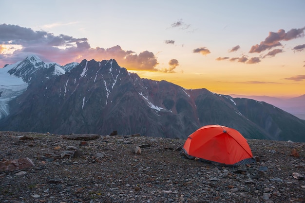 Scenic alpine landscape with tent at very high altitude with view to large mountains in orange dawn sky Vivid orange tent with awesome view to high mountain range under cloudy sky in sunset colors