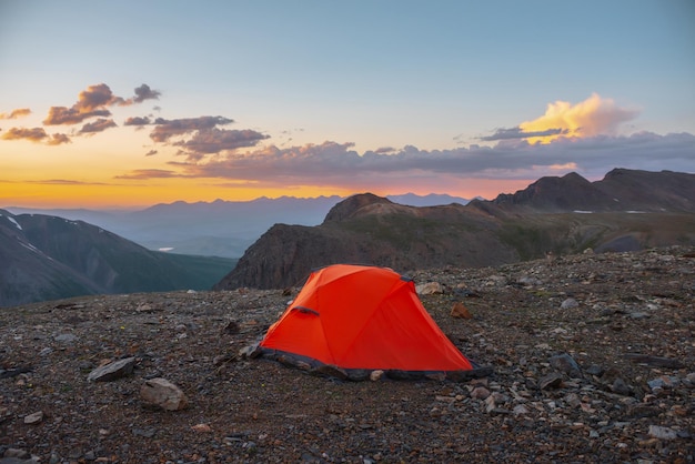 Scenic alpine landscape with tent at very high altitude with view to large mountains in orange dawn sky Vivid orange tent with awesome view to high mountain range under cloudy sky in sunset colors