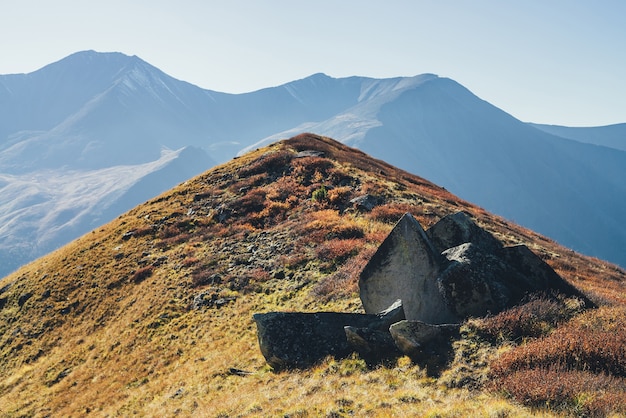 Scenic autumn landscape with sharp shattered stone on red hill on background of mountains silhouettes in sunlight. Colorful mountain scenery with hill in fall colors. Wonderful autumn alpine view.