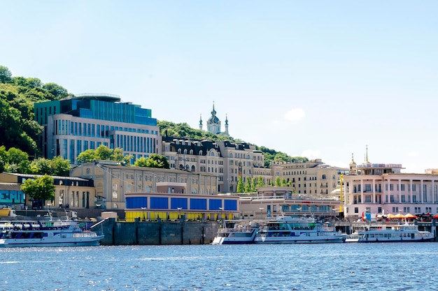 Scenic bright travel cityscape of river Dnipro in KievUkraine with blue water bridgepleasure shipsresidential buildings in river port Andrews church at sunny summer dayKiev Ukraine11 June 2021