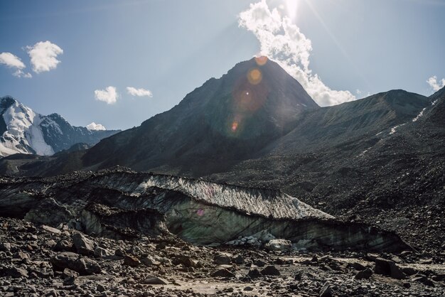 Scenic highlands landscape with big cracked glacier with scratches among moraines on background of high mountain peak in sunlight. Awesome mountain scenery with large glacier and great mountain top.