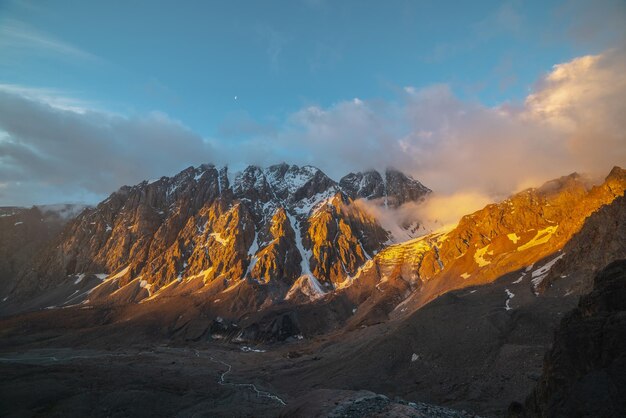 Scenic landscape with snowy mountain wall and vertical glacier with icefall in low clouds in golden sunrise colors Colorful view to snow mountains and rocks in gold morning sunlight in low clouds