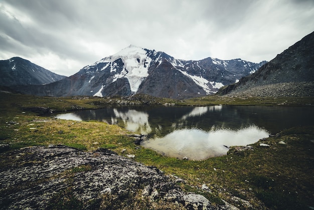 Scenic mountain landscape with glacial lake among mountains and glacier under gray cloudy sky. Atmospheric alpine scenery with transparent mountain lake in rainy weather. Beautiful clear alpine lake.