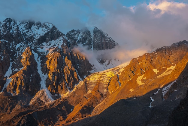 写真 黄金の日の出の色の低い雲の中に雪に覆われた山頂と氷河がある風光明媚な山の風景 低い雲の中で金色の朝の日差しの中で氷瀑のある垂直氷河にカラフルな山の景色