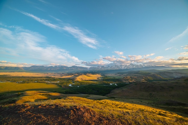 Scenic sunny view from sunlit grassy hill to forest valley with serpentine river against high snowy mountain range in sunlight Beautiful snake mountain river in forest and snow mountains in sunshine