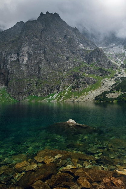 Scenic view of foggy mountains cover by dark clouds and green forest with a reflection in a lake Stony shore Morskie Oko Marine Eye High Tatras Zakopane Poland