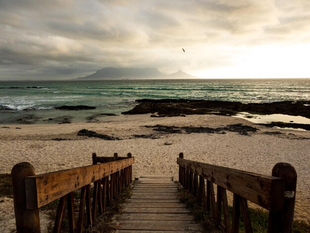 Photo scenic view of a wooden walkway leading down to a cloudy beach with table mountain in the distance