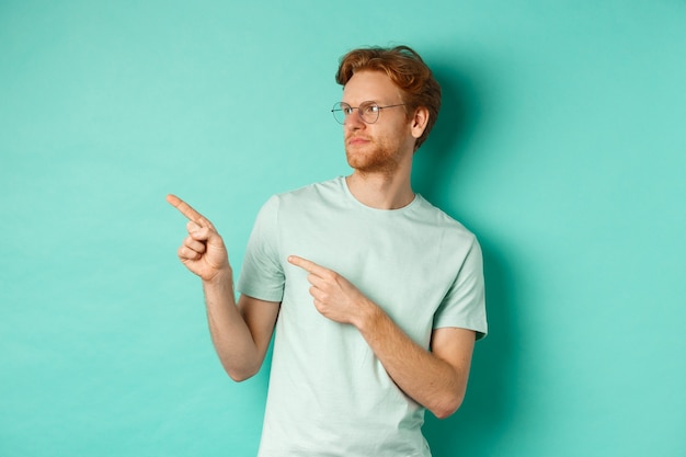 Sceptische blanke man met rood haar en baard, met bril en t-shirt, kijkend en wijzend teleurgesteld naar links, iets slechts beoordelend, staande over mint achtergrond