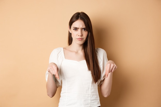 Sceptische jonge vrouw fronst, handen naar beneden wijzend op iets slechts of teleurstellend, kijkt boos, staande op beige achtergrond.