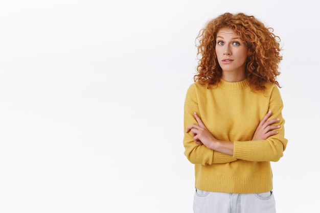 Sceptische mooie roodharige vrouw in gele trui, met krullend haar, kruisarmen op de borst, wenkbrauwen optrekken en staren met ongeloof, luister naar iemand die liegt, kijk veroordelend over de witte muur