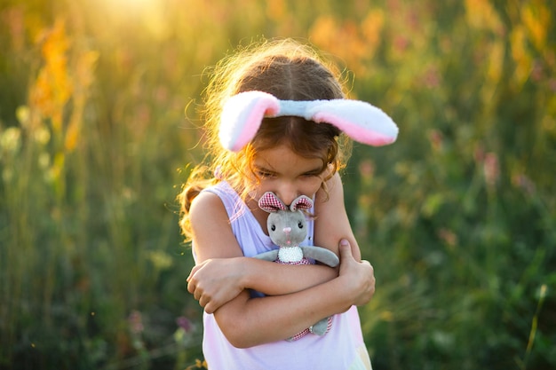 Schattig 5-jarig meisje met konijnenoren knuffelt zachtjes een speelgoedkonijn in de natuur in een bloeiend veld in de zomer met gouden zonlicht. Pasen, paashaas, jeugd, gelukkig kind, lente.