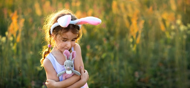 Schattig 5-jarig meisje met konijnenoren knuffelt zachtjes een speelgoedkonijn in de natuur in een bloeiend veld in de zomer met gouden zonlicht. Pasen, paashaas, jeugd, gelukkig kind, lente.