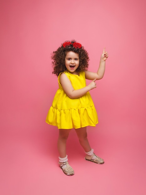 Schattig aantrekkelijk vrolijk meisje in gele jurk wijzend met haar wijsvinger uppink pastelkleurige achtergrond