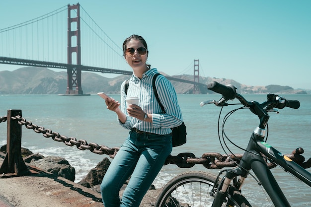 Schattig Aziatisch meisje in zonnebril geniet van mobiele telefoon op het strand tijdens een fietstocht. jonge vrouw met koffie en smartphone zittend op ijzeren touw richting zoeken op online kaart. golven in de oceaan in de zomer
