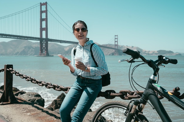 Schattig aziatisch meisje in zonnebril geniet van mobiele telefoon op het strand tijdens een fietstocht. jonge vrouw met koffie en smartphone zittend op ijzeren touw richting zoeken op online kaart. golven in de oceaan in de zomer