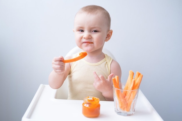 schattig babymeisje in gele bodysuit zittend in de stoel van het kind wortelschijfjes eten