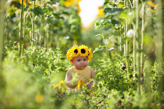 Schattig babymeisje met een zonnebloemkrans zittend op het zonnebloemveld en onderzocht de natuur rondom